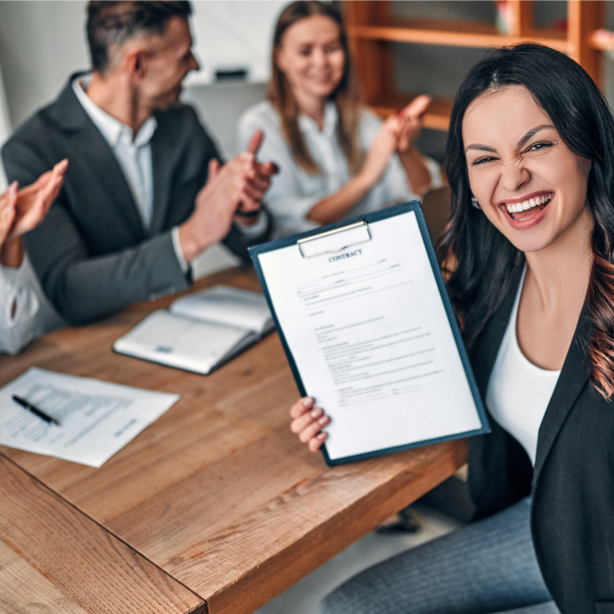 Fröhliche Personen an einem Tisch. Die Frau im Vordergrund hält einen unterschriebenen Arbeitsvertrag in der Hand. 